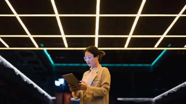 一位女子盯着手中的平板电脑，背景是被灯光分割的暗室天花板