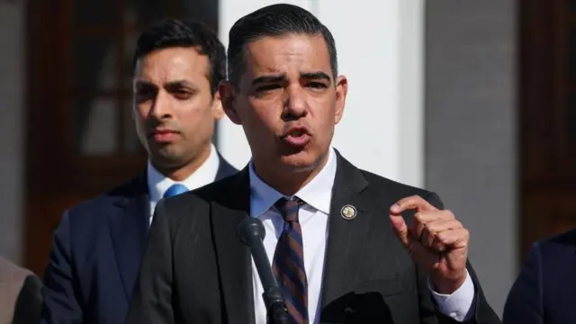 白い建物を背に、ダークスーツ姿の男性が演台のマイクを前に話している