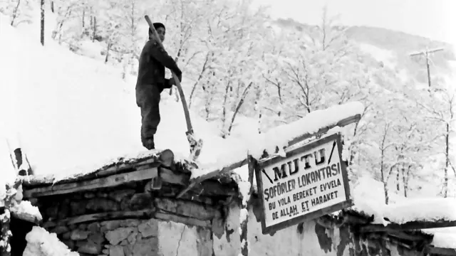 Bir çocuk, elindeki kürekle, karla kaplı tek katlı bir yapının üzerindeki karları temizliyor. Yapının üstünde bir tabela var. Tabelada, "Mutu Şoförler Lokantası: By yola bereket evvela, de Allah et hareket" yazıyor. 