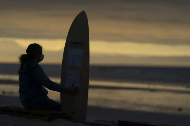 A woman stands with a surfboard on the beach in the twilight.