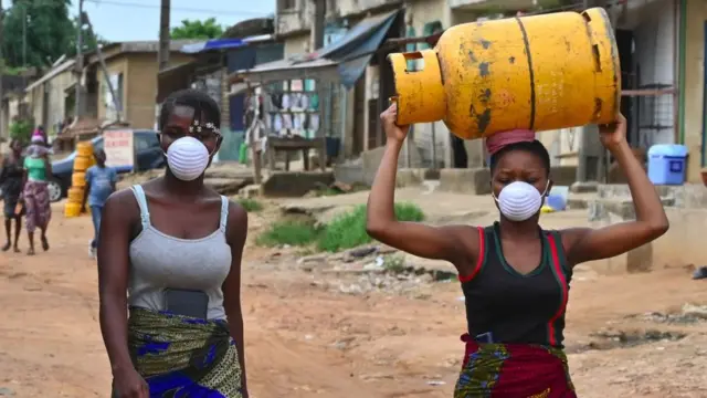 Duas mulheres negras usando máscara enquanto caminham na rua, uma delas carregando um botijãocs roletagás na cabeça