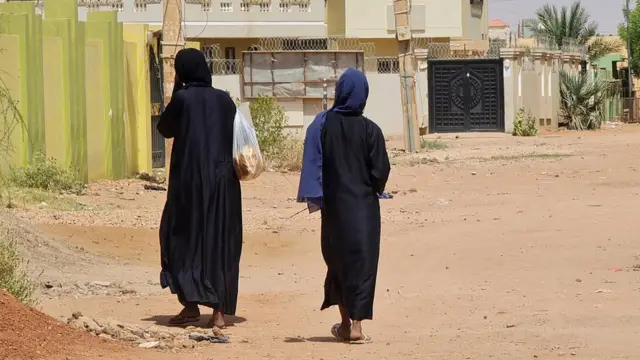 Deux femmes vêtues de manteaux noirs se dirigent vers des immeubles résidentiels, dos à la caméra. L'une d'elles porte un sac rempli de pain.