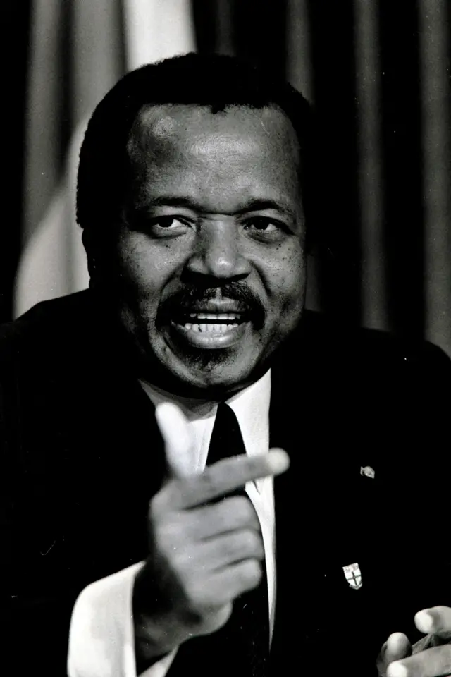 Fotografia em preto e branco do presidente dos Camarões, Paul Biya, com aparência mais jovem, vestindo um terno e fazendo um gesto com a mão direita