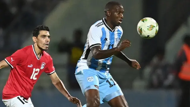 Tumisang Orebonye (Botwana), vêtu d'un maillot rayé bleu clair et blanc, tente de contrôler un ballon de football sous le regard du défenseur égyptien Mohamed Hamdy.