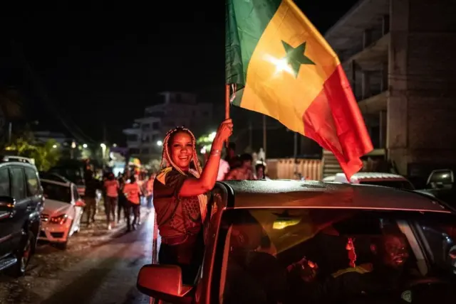 Une femme se penche par la fenêtre d'une voiture et brandit le drapeau sénégalais.