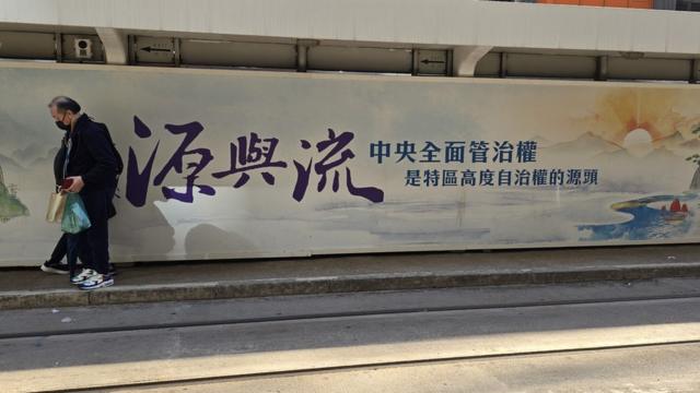 香港北角某电车站一位乘客走过宣传中国对香港“全面管治权”的大型广告（BBC中文图片12/3/2024）