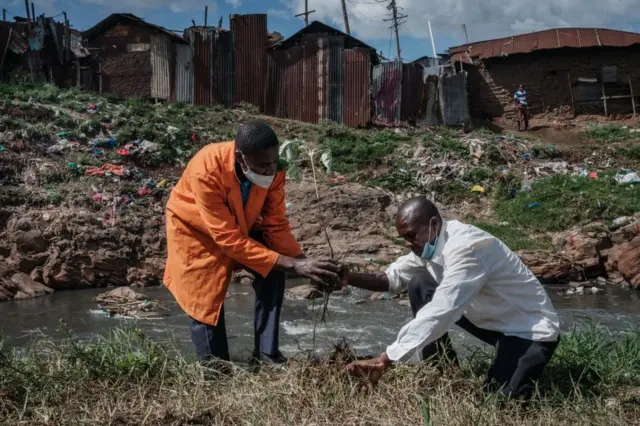 Two men plant a tree on a littered riverbank.