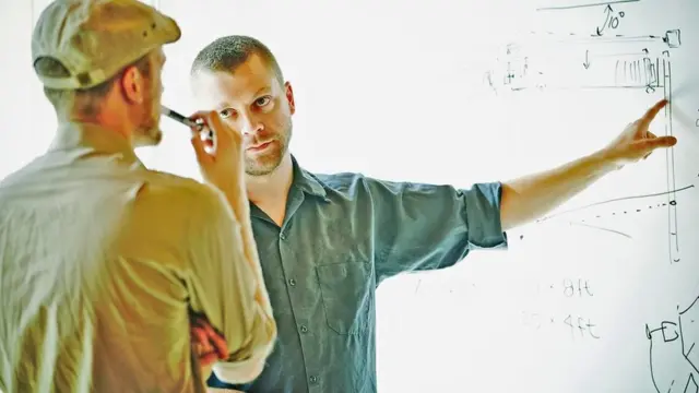 Una persona dándole instrucciones a otra sobre un dibujo en una pizarra