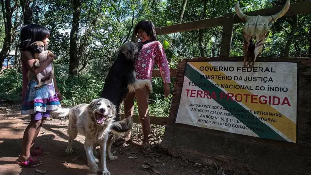 Dos pequeñas indígenas juegan con sus perros frente a un letrero de la FUNAI que anuncia la protección de un terreno guaraní en Brasil