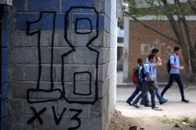 Pintada del Barrio 18 en una pared y al fondo escolares.