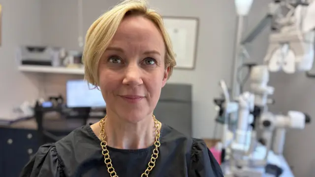Une femme blonde aux cheveux coupés au carré. Elle porte un haut noir et un collier à chaîne dorée. Elle se trouve dans une pièce d'un cabinet d'opticien.