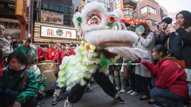 Pessoa vestida de dragão na rua, em frente a comércio, observada por público