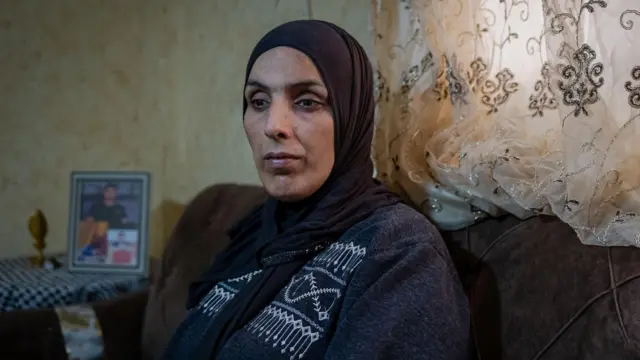 Zbliżenie na matkę Jada, wyglądającą na opuszczoną, siedzącą w salonie rodzinnym w obozie dla uchodźców Al-Far'a na okupowanym Zachodnim Brzegu. Za nią na stole stoi oprawione zdjęcie Jada.