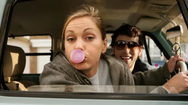 Хлопець і дівчина в авто. Він сміється, вона надуває бульбашку з жувальної гумки