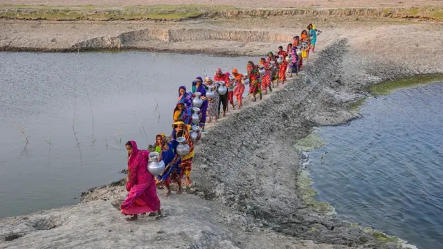 Une file de femmes vêtues de vêtements colorés et coiffées de foulards, portant chacune un grand pot gris, marchent vers la caméra le long d'une étroite berge surélevée entre deux grands bassins d'eau.