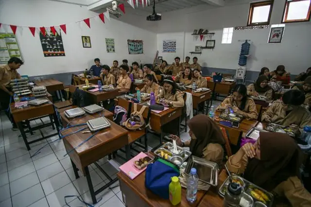 Sekelompok siswa makan makanan bergizi gratis (MBG) di kelas mereka di Sekolah Menengah Atas Negeri 20 Bandung, Jawa Barat, Indonesia, pada tanggal 14 April 2025. Dadan Hindayana, Kepala Badan Gizi Nasional Indonesia (BGN), menyatakan bahwa pada akhir tahun 2025, program MBG diharapkan mencapai 82,9 juta penerima manfaat.