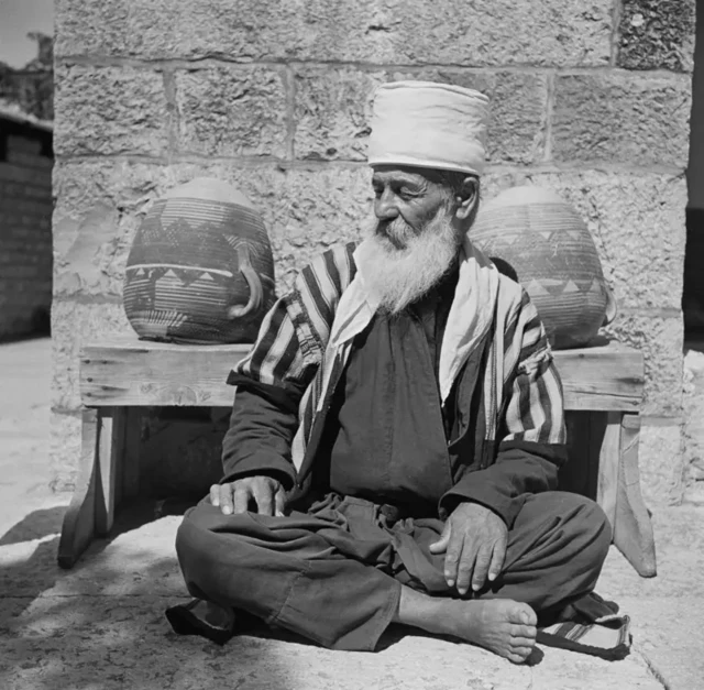 عکس قدیمی سیاه و سفید، یکی از شیوخ لبنانی دروزی از مرتبه دوم، با ریش بلند سفید، عبای سفید و سیاه راهراه در حاصبیای لبنان، روی زمین چهارزانو نشسته، پشت سر او دو کوزه روی یک نیمکت چوبی کوچک