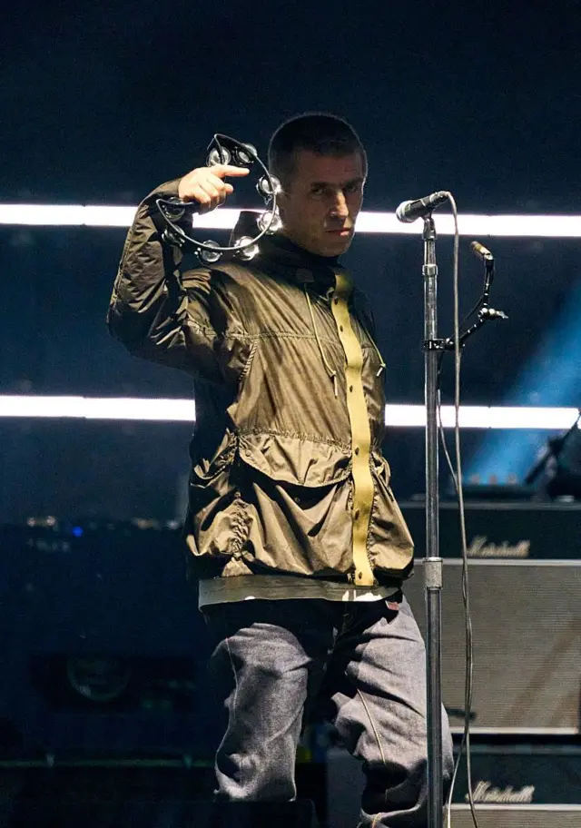 Liam Gallagher é fotografado em frente a um microfone no palco, segurando um pandeiro. Ele está vestindo uma jaqueta verde.