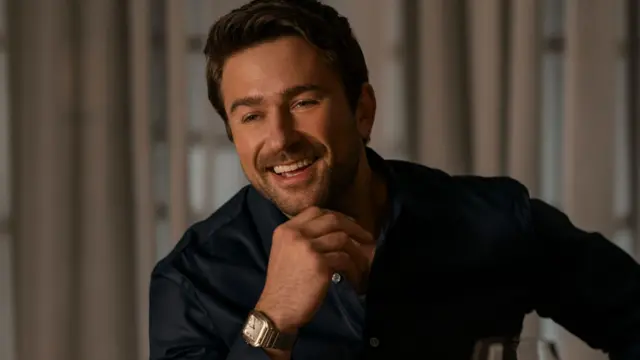 O ator Brandon Sklenar em cena no filme "A Empregada". Ele está sorrindo enquanto apoia o queixo na mão e veste uma camisa de cor escura