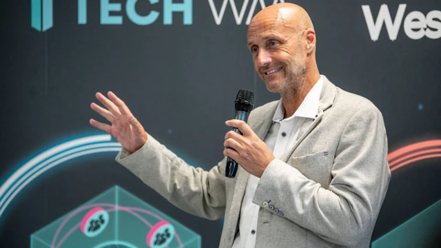 Andy Hague viste un traje color crema y una camisa blanca. Sostiene un micrófono con la mano izquierda y mueve la derecha al dirigirse a la audiencia. Está de pie frente a carteles promocionales de TechWM. Tiene la cabeza calva y una barba corta.