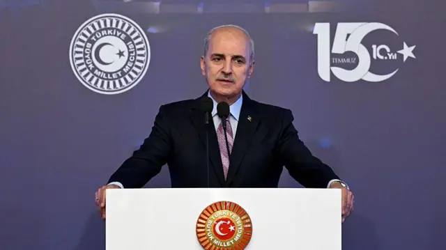 Numan Kurtulmuş ellerini kürsünün iki yanına koymuş konuşma yapıyor. Önünde TBMM forsu amblemi var. Kürsü beyaz.