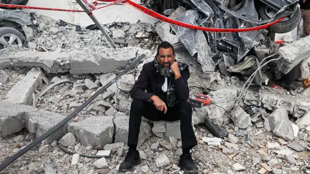 Man sitting beneath wrecked car looking at the camera with wrecked car behind him