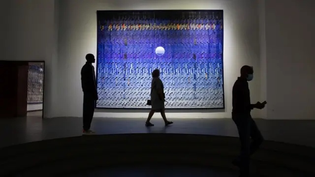 Des spectateurs passent devant une œuvre de l'artiste malien Abdoulaye Konaté à l'Ancien Palais de Justice, le 21 mai 2022, dans le cadre de la Biennale de Dakar. L'œuvre d'art présente différentes nuances de bleu et contient ce qui ressemble à la lune.