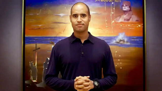 سيف الإسلام القذافي عام 2002 ، يقف أمام لوحة تحمل صورة والده معمر القذافي في الزاوية اليمنى العليا.