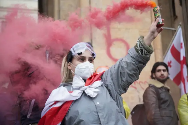 Девушка в маске и с дымовой шашкой откуда валит розовый дым.