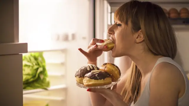 Mujer comiendo dulces en frente de la nevera.