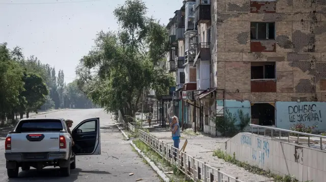 One resident bin dey stand on top di street outside one apartment – wey im dey tok wit one police officer wey comot im head from di police van for Toretsk near di frontline for di Donetsk region in July 2024. 