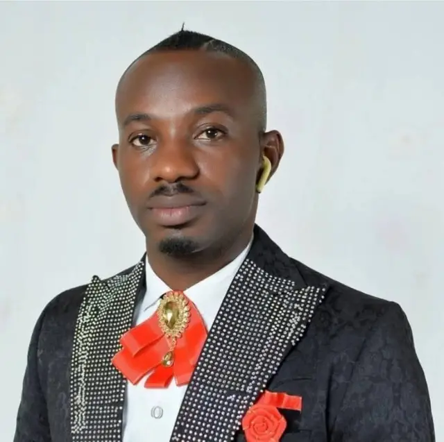 Human Right Activist, Comrade Israel Joe wear blaxk suit wit orange clour bow tie and pocket fillers. E wear wgite sgirt insite di suit wey get silver and gold stone decisions for di from open collar.