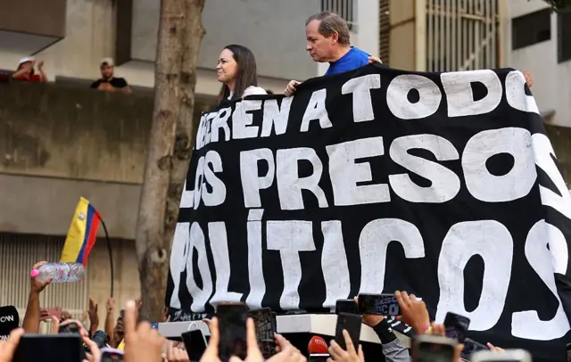 María Corina Machado y Juan Pablo Guanipa sobre un camión sostienen un cartel donde se lee "Liberen a todos los presos políticos". 