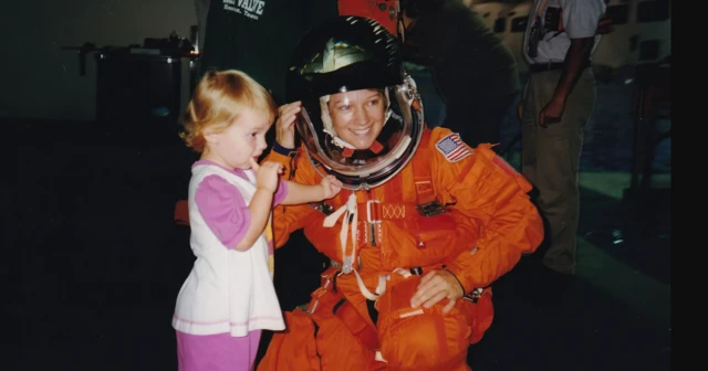 अन्तरिक्षयात्री आइलीन कोलिन्सले सुन्तला रङ्गको स्पेससूट र पारदर्शी हेलमेट लगाएकी छिन्। उनी तीन वर्षकी छोरीको छेउमा झुकेर मुस्कुराउँछिन्।
