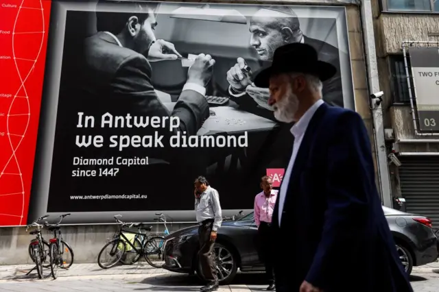 Escena urbana en Amberes con varias personas caminando frente a un gran cartel publicitario que dice: ‘In Antwerp we speak diamond – Diamond Capital since 1447’ ("En Amberes hablamos el idioma de los diamantes: Capital mundial del diamante desde 1447"). Muestra una imagen en blanco y negro de dos personas examinando diamantes. Hay bicicletas estacionadas junto a la acera y un automóvil estacionado frente al cartel.