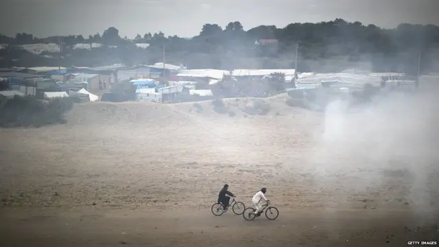 Migrants cycle through smoke from a fire burning rubbish at the Jungle migrant camp on September 6, 2016 in Calais, France.