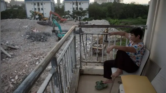 This file photo taken on October 12, 2016 shows a woman sitting on her balcony as she watches bulldozers clearing the rubble of demolished buildings to clear the ground for a new property development project in Sanya, on China"s southern Hainan island