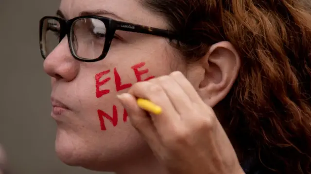 Mujer a la que le escriben en la mejilla las palabras "EleNão".