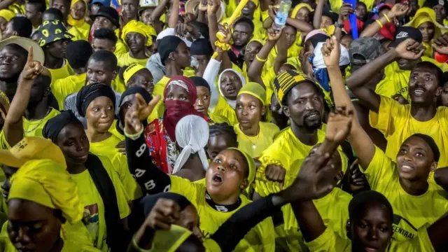 Dandazon mutane sanye da tufafi ruwan dorawa. Akwai wata mace a tsakiya sanye da tufafi launin jinin kare, rufe da fuskarta idanunta na nuna kwarin gwiwa.