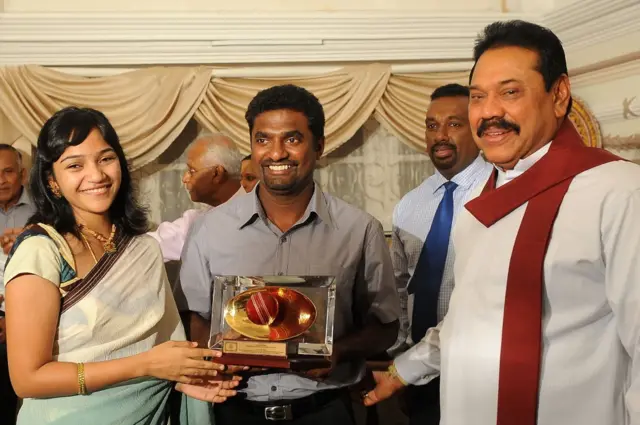 Sri Lankan President Mahinda Rajapakse (R) presents a gift to star cricketer Muttiah Muralitharan (C) flanked by his wife Madhi Malar (L) during a tea party at the official Presidential residency in Colombo on April 4, 2011. Sri Lanka lost to India by six wickets in a thrilling World Cup final on April 2, in Mumbai. AFP PHOTO / Lakruwan WANNIARACHCHI (Photo credit should read LAKRUWAN WANNIARACHCHI/AFP via Getty Images)