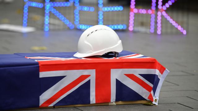 伦敦一场声援香港集会上摆放盖上旧英属香港旗帜与白色安全帽的假棺材（12/12/2020）