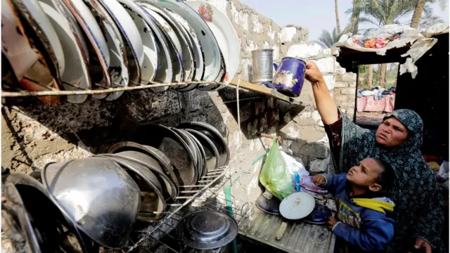 A woman placing her kitchen utensils to dry outside. There are plates lined up on a shelf to her right.