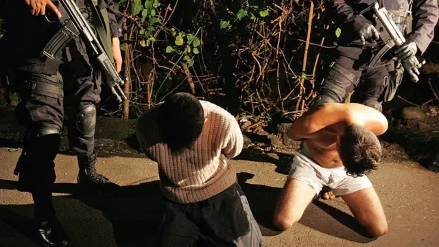 Dos hombres arrodillados bajo custodia de dos agentes de la policía