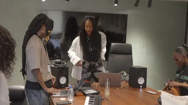 Tems regarde une jeune femme qui utilise un ordinateur portable connecté à divers équipements musicaux.