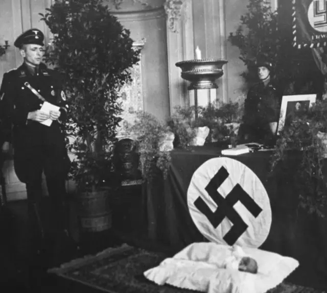 Oficial nazista olha para um criança deitadazambia 1xbetberçozambia 1xbetfrente a uma bandeira com suástica