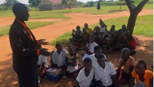 Mwalimu Mkuu wa shule ya msingi ya Bukedea Okurut Levy(pichani mbele ya wanafunzi ) anasema imekuwa ni changamoto kubwa kuwafundisha watoto kando ya barabara