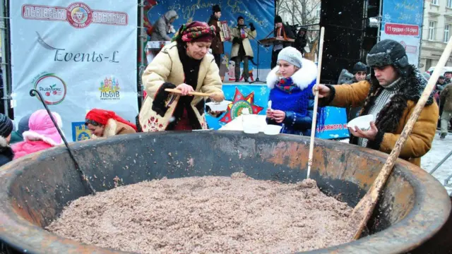 У величезному казані вариться різдвяна кутя, біля казана стоять  жінка і чоловік у національному вбранні, а також відвідувачі. 