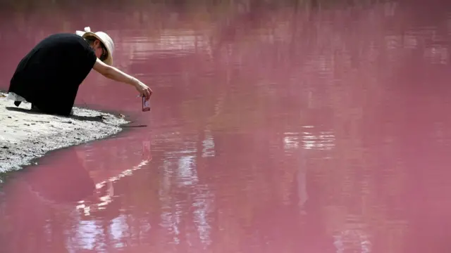 Uma mulher se agacha perto da borda do lago rosa para tirar uma foto