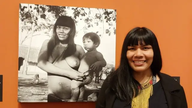 Fotografia colorida mostra uma mulher indígenacomo sacar o bonus da arbetycabelo preto comprido e franja ao ladocomo sacar o bonus da arbetyuma fotografia preto e branca na parede na qual uma mulher indígena segura um bebê no colo