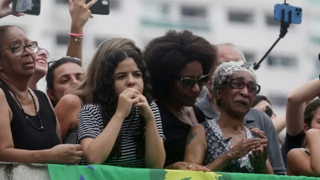 Fotografia colorida mostra pessoas chorando; no canto à direita há uma senhora idosa negracasino online leovegascabelo branco, com faiza no cabelo e óculos